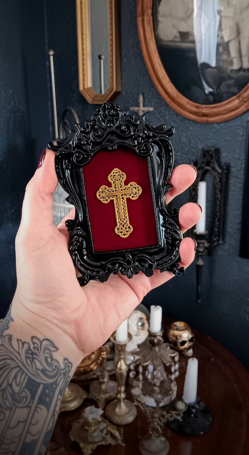 May include: A black ornate frame with a red velvet mount and a gold cross inside. The frame is held in a hand with a tattoo.