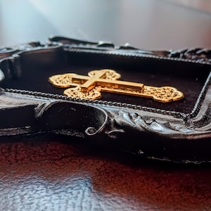 May include: A gold cross with intricate details is displayed in a black ornate frame. The frame has a velvet backing and a distressed finish.