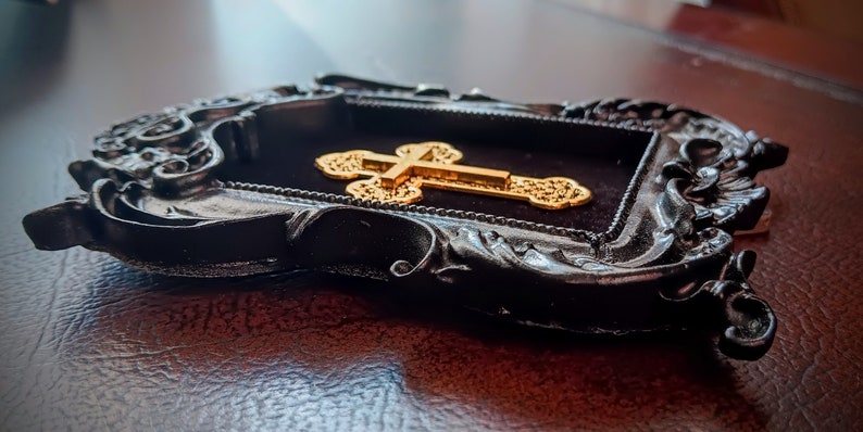 May include: A gold cross with intricate details is displayed in a black ornate frame. The frame has a velvet backing and a distressed finish.