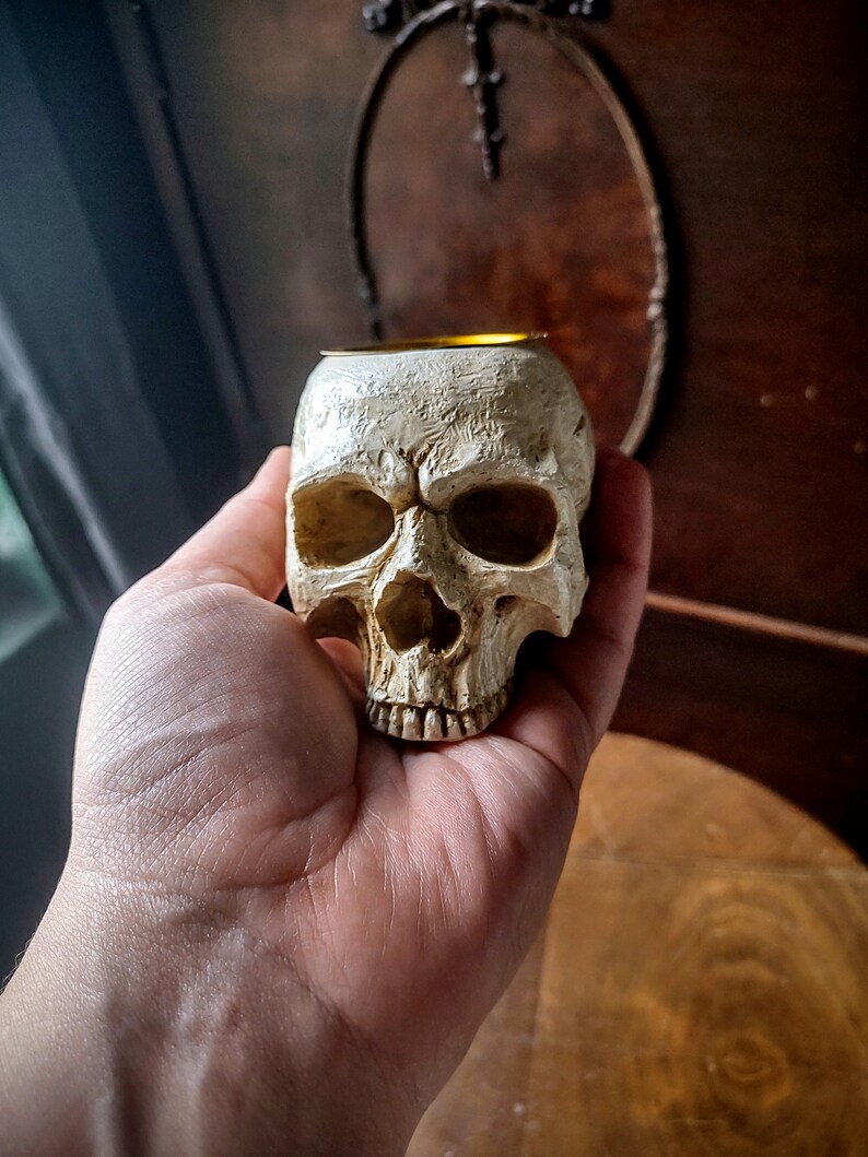 Pode incluir: Um porta-velas de cer&acirc;mica branca em forma de cr&acirc;nio com uma borda dourada. O cr&acirc;nio &eacute; detalhado com caracter&iacute;sticas realistas, incluindo &oacute;rbitas oculares, um nariz e dentes.