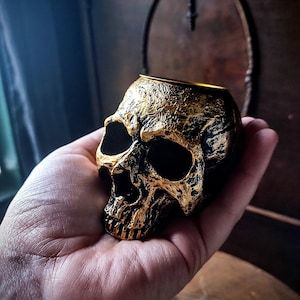 Pode incluir: Um porta-velas de caveira dourado com interior preto. A caveira &eacute; detalhada com uma superf&iacute;cie texturizada e uma parte superior oca para uma vela.