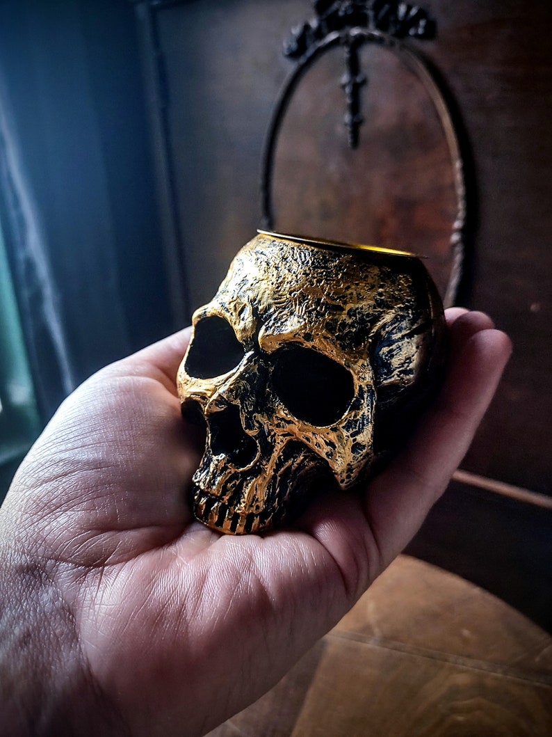 Pode incluir: Um porta-velas de caveira dourado com interior preto. A caveira &eacute; detalhada com uma superf&iacute;cie texturizada e uma parte superior oca para uma vela.