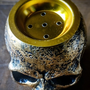 Pode incluir: Um pote de cachimbo de &aacute;gua em forma de caveira dourada com uma parte superior redonda de cor dourada e cinco pequenos orif&iacute;cios. A caveira &eacute; detalhada com uma superf&iacute;cie texturizada.