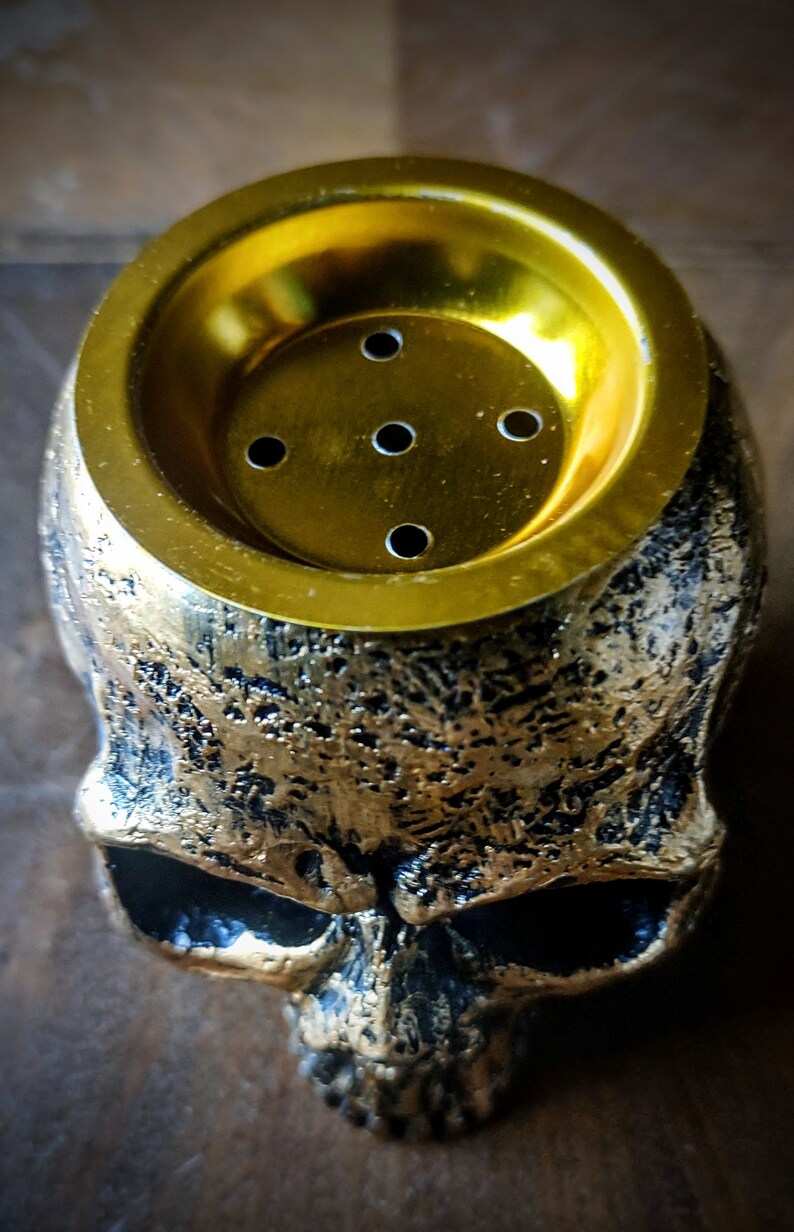 Pode incluir: Um pote de cachimbo de &aacute;gua em forma de caveira dourada com uma parte superior redonda de cor dourada e cinco pequenos orif&iacute;cios. A caveira &eacute; detalhada com uma superf&iacute;cie texturizada.