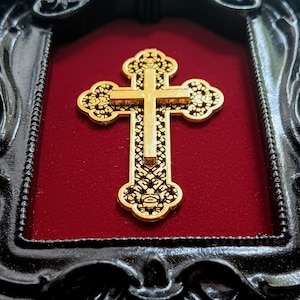 May include: A gold-coloured cross with intricate details is displayed in a black ornate frame with a red velvet background.