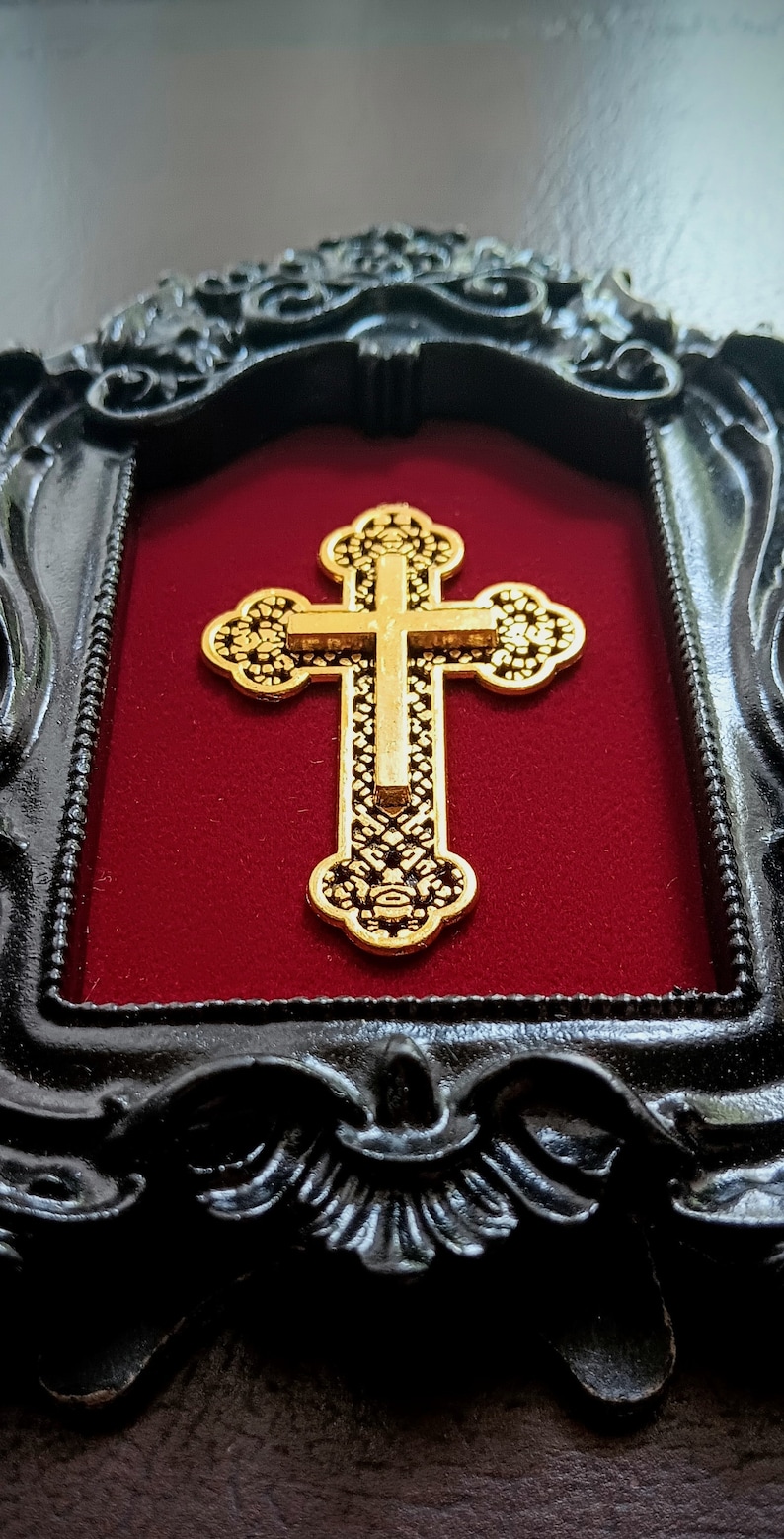 May include: A gold-coloured cross with intricate details is displayed in a black ornate frame with a red velvet background.