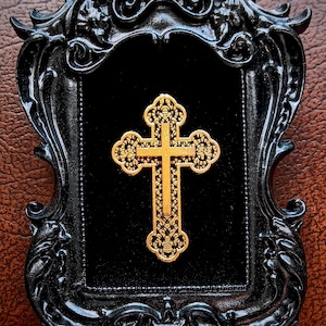 May include: A gold cross with intricate details is displayed in a black ornate frame. The frame has a velvet backing.