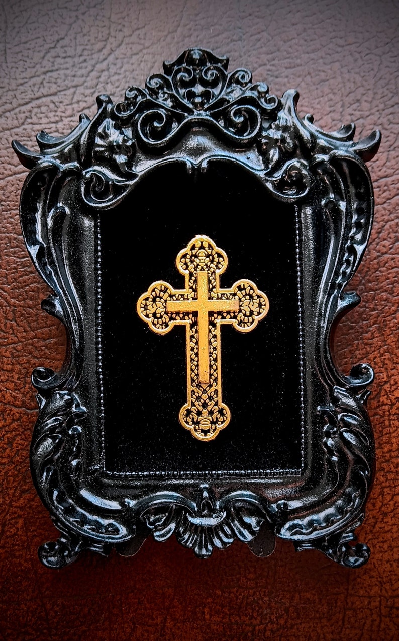 May include: A gold cross with intricate details is displayed in a black ornate frame. The frame has a velvet backing.