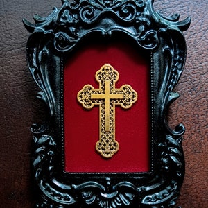 May include: A gold-coloured cross with intricate details is displayed in a black ornate frame with a red velvet background.