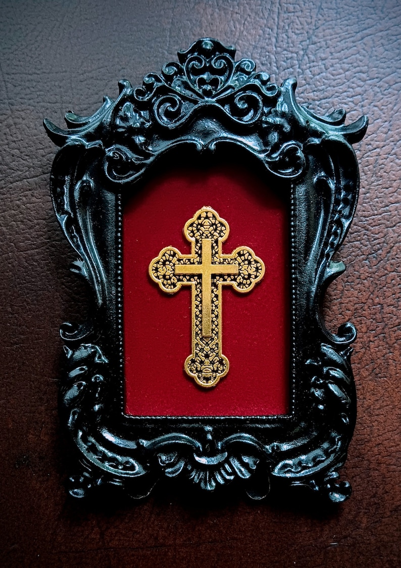 May include: A gold-coloured cross with intricate details is displayed in a black ornate frame with a red velvet background.