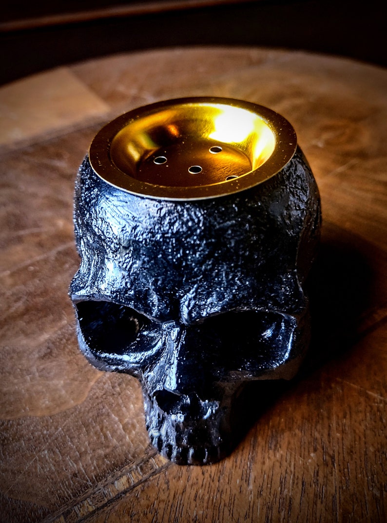 Pode incluir: Queimador de incenso preto em forma de caveira com uma parte superior de metal dourado e furos para queimar incenso.