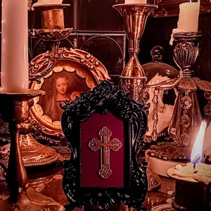 May include: A black framed picture with a gold cross on a red background. The picture is surrounded by candlesticks with lit candles and other decorative items.