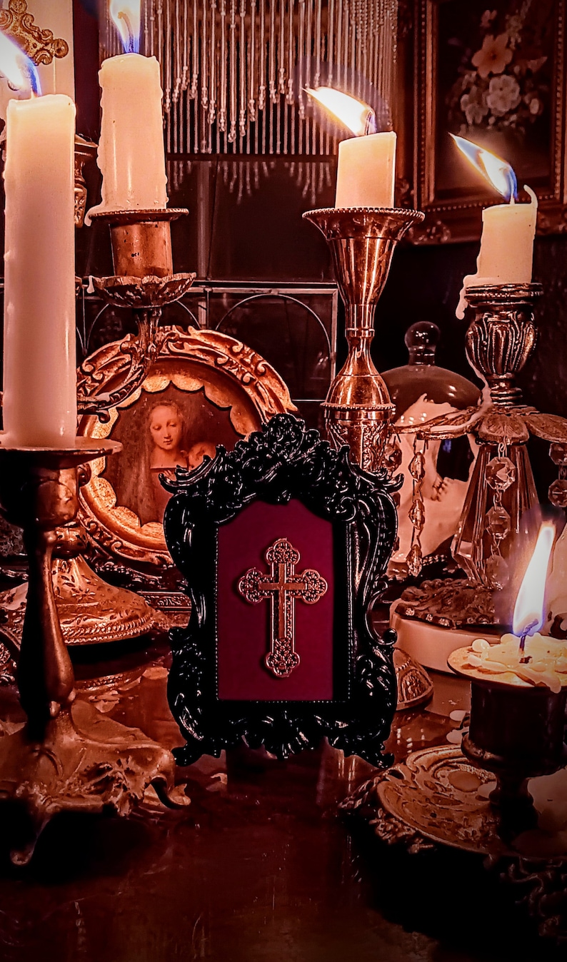 May include: A black framed picture with a gold cross on a red background. The picture is surrounded by candlesticks with lit candles and other decorative items.