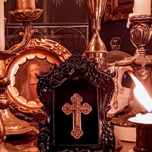 May include: A black ornate frame with a gold cross inside. The frame is sitting on a table with candles and other decorative items.