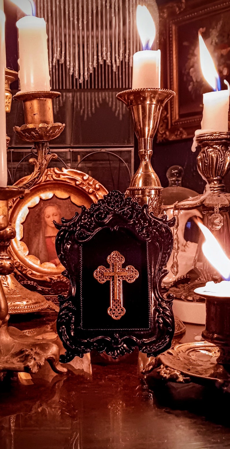 May include: A black ornate frame with a gold cross inside. The frame is sitting on a table with candles and other decorative items.