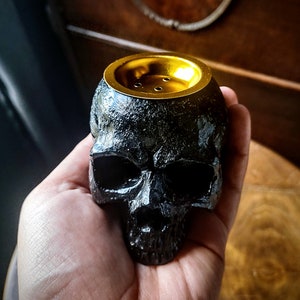 Pode incluir: Queimador de incenso preto em forma de caveira com topo dourado. O queimador tem uma superf&iacute;cie texturizada e um pequeno furo no topo para o incenso.