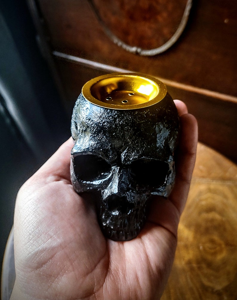 Pode incluir: Queimador de incenso preto em forma de caveira com topo dourado. O queimador tem uma superf&iacute;cie texturizada e um pequeno furo no topo para o incenso.
