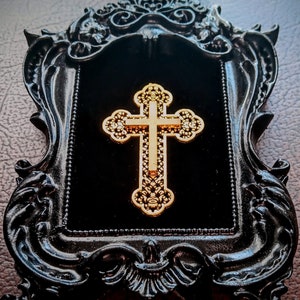 May include: A gold cross with intricate details is displayed in a black ornate frame with a velvet backing.