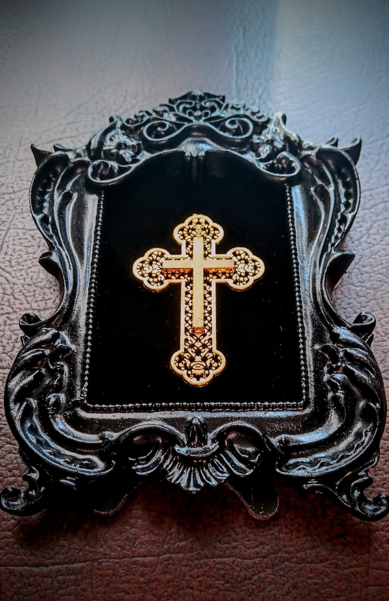 May include: A gold cross with intricate details is displayed in a black ornate frame with a velvet backing.