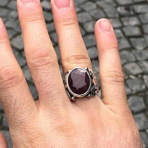 Può includere: Anello in argento con una grande pietra preziosa rosso scuro a taglio ovale. L'anello presenta dettagli intricati attorno alla pietra e una fascia color oro. L'anello è su una mano, con una manica di giacca di jeans sullo sfondo.
