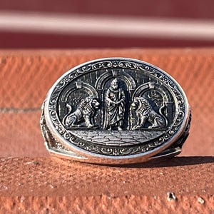 May include: An oval silver ring featuring a detailed relief carving of a central standing figure flanked by two lions. The design is set within an arched architectural frame, with intricate decorative patterns along the outer edge of the ring face.