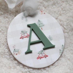 May include: A circular Christmas decoration with a green letter "A" on a grey and white checked pattern. The design includes illustrations of red cars with Christmas trees, houses, and fir trees. A white ribbon is attached.