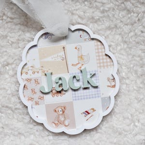 May include: A round, scalloped-edge baby name plaque with the name "Jack" in light green letters. The plaque features a patchwork design with illustrations of toys, a goose, and a teddy bear in soft pastel colors.