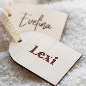 以下が含まれることがあります： 名前が書かれた木製のギフトタグが2つ。上のタグには「Evefina」と書いてあり、下のタグには「Lexi」と書いてあります。