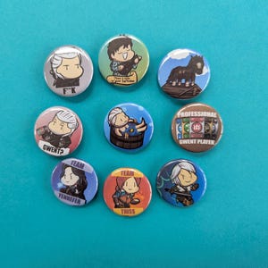 May include: A collection of nine round pinback buttons with cartoon illustrations. The buttons feature characters and text, including "F*K", "GWENT?", "TEAM YENNEFER", "TEAM TRISS", and "PROFESSIONAL GWENT PLAYER". The buttons are on a teal surface.