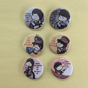 May include: Six round pinback buttons with cartoon illustrations and text. The buttons feature images of a person playing an instrument, and other illustrations with text that reads "Northern Downpour Sends Its Love", and "It's Nine in the Afternoon".