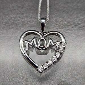 May include: A silver pendant necklace featuring a heart-shaped design with the word "MOM" in the center. The pendant is adorned with a row of small, sparkling gemstones along one side, and hangs from a delicate chain.