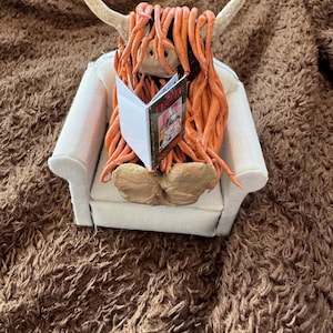 May include: A novelty Highland cow figurine with orange hair and horns, reading a miniature book while seated in a small white armchair. The cow is resting on a brown, textured surface.