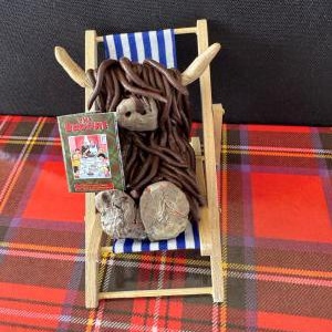 May include: A miniature Highland cow figurine with brown fur and horns, relaxing in a wooden deckchair with a blue and white striped seat. The cow is holding a small book with colorful illustrations. The chair is on a red tartan blanket.