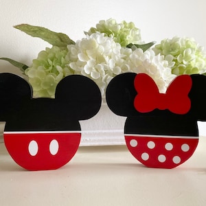 May include: A pair of wooden Mickey Mouse and Minnie Mouse head ornaments. The Mickey Mouse ornament is black with red ears and a red face with two white circles. The Minnie Mouse ornament is black with red ears and a red bow. Both ornaments have white bead garlands.