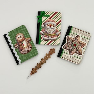 May include: A collection of Christmas-themed notebooks and a pen. The notebooks feature festive designs including a gingerbread man, Santa Claus, and a star-shaped cookie. The pen is adorned with small gingerbread man figures.