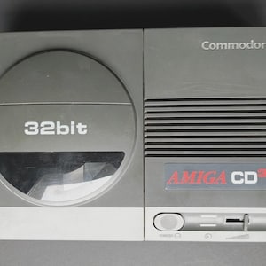 Puede incluir: Una consola gris Commodore Amiga CD32. La consola presenta una unidad de disco circular, el texto "32bit" y el logotipo de Amiga CD32. La consola tiene forma rectangular con una base blanca.