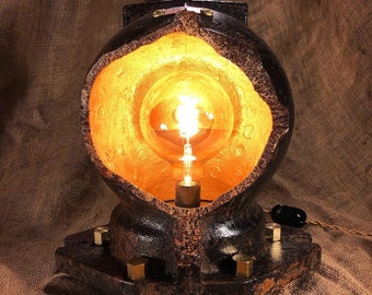 Puede incluir: Una lámpara de mesa de estilo steampunk hecha a partir de un accesorio de tubería industrial reutilizado. La lámpara tiene un interior pintado de oro y una bombilla de vidrio transparente. La base es de metal oscuro con un acabado oxidado.