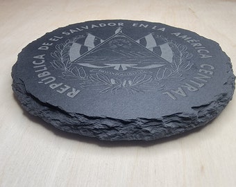 May include: A round, dark gray slate coaster with engraved text and a central emblem. The emblem features a triangle, flags, and text, with the words "REPUBLICA DE EL SALVADOR EN LA AMERICA CENTRAL" around the edge.