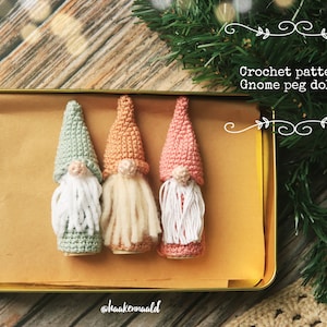 Op de afbeelding: Drie gehaakte dwerg-steekpoppen in een gouden blik. De dwergen zijn groen, perzikkleurig en roze met witte garenbaarden. De tekst "Crochet pattern Gnome peg dolls" staat boven de blik.