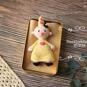 Puede incluir: Una muñeca de crochet con un vestido amarillo, un corazón rosa y pelo negro. La muñeca está sentada en una caja de hojalata sobre un fondo de madera marrón. El texto "Haakpatroon BUMBA" es visible en la imagen.