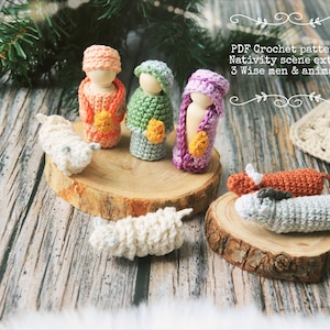 Może przedstawiać: Scena Bożego Narodzenia na szydełku z trzema mędrców, dwoma owcami i krową. Figurki są wykonane z włóczki i mają drewniane kołki jako ciała. Scena jest ustawiona na drewnianej powierzchni.