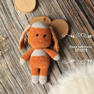 Op de afbeelding: Een gehaakt oranje en wit pluche speelgoed in de vorm van een hond. Het speelgoed ligt op een houten oppervlak met een wit bontkleed op de achtergrond. De tekst "Haakpatroon DROPJE" is zichtbaar in de afbeelding.