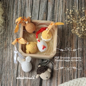 Op de afbeelding: Een set van vijf gehaakte paaseieren dieren in een houten doos. De dieren zijn een konijn, een kuiken, een haan, een lam en een konijn. De dieren zijn allemaal in pastelkleuren en hebben schattige gezichten. De doos is gevuld met stro en heeft een rood lint eromheen gebonden. De tekst "PDF Crochet pattern Easter eggs animals" staat op het houten oppervlak.