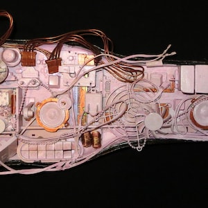 May include: A pink guitar-shaped object decorated with various small objects, including metal, plastic, and fabric. The object is covered in a layer of white paint, giving it a vintage look.