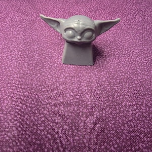 May include: A gray mechanical keyboard keycap featuring a sculpted figure with large oval eyes, a small nose, and wide pointed ears. The base of the keycap is a square shape designed to fit onto a mechanical switch stem, with a smooth matte finish.