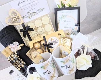 May include: A gift basket filled with items for a wedding or engagement celebration. The basket includes two mugs with the words "Mr." and "Mrs." on them, a box of chocolates, candles, a black and white blanket, and a bottle of body spray. The basket is decorated with white and gold flowers.