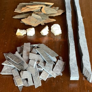 May include: A collection of slate tiles in various shapes and sizes, including rectangular, square, and irregular shapes. The tiles are a dark gray color and have a rough, textured surface. There are also some white, irregularly shaped stones.