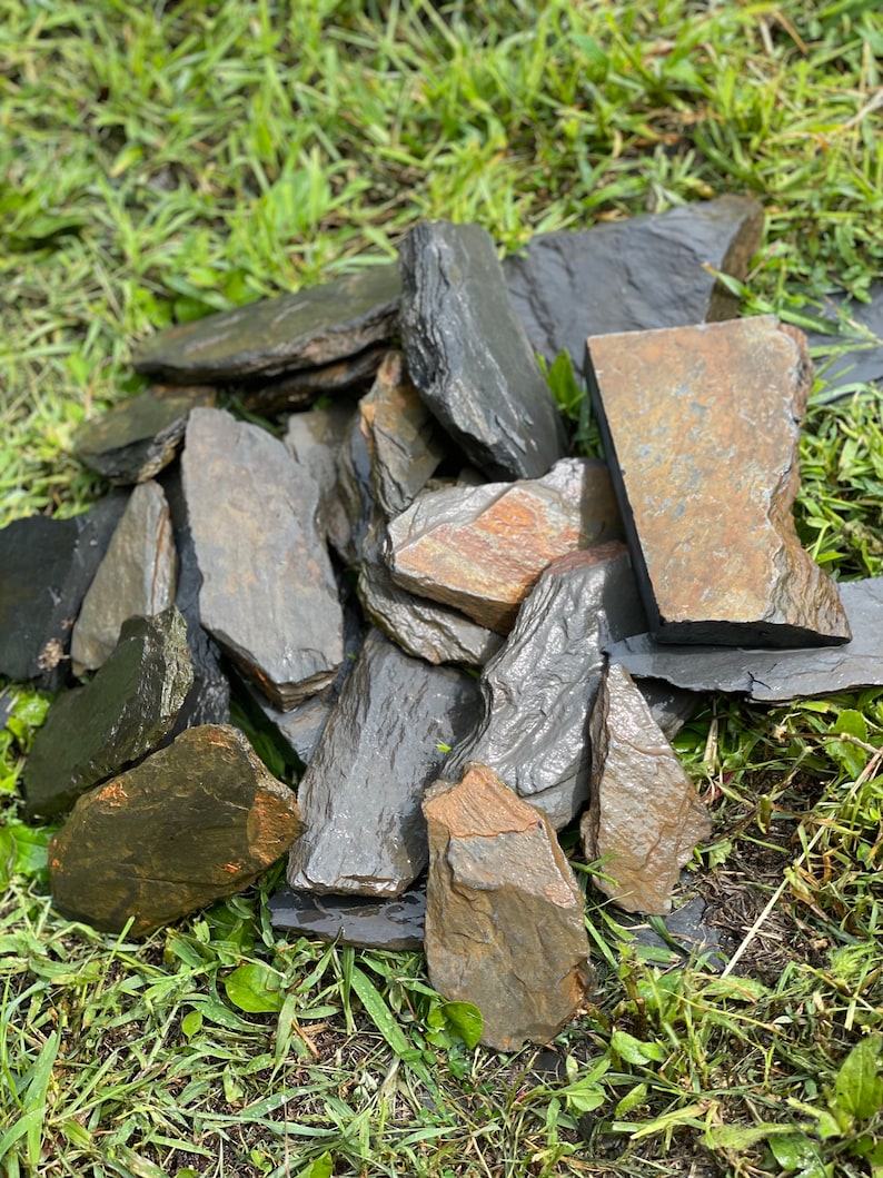 以下が含まれることがあります： 緑の草の上に、暗い灰色と茶色の粘板岩の石が積み重ねられています。石のいくつかは、ざらざらした質感です。