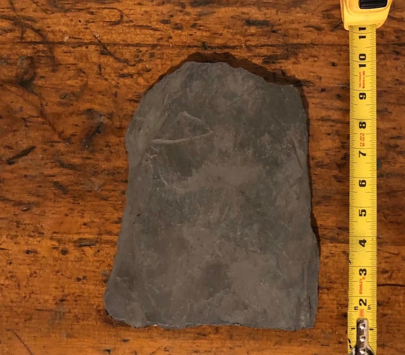 May include: A dark gray slate tile measuring approximately 6 inches by 8 inches. The tile is smooth and has a slightly rough texture.