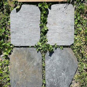 May include: Four slate tiles in various shapes and sizes, with a light gray color and a rough texture. The tiles are arranged in a square pattern on a bed of green grass.